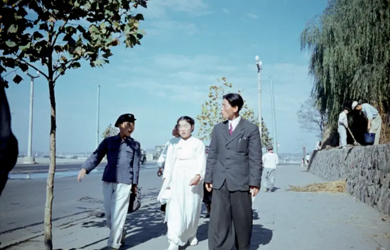 Eine Gruppe von drei Menschen, gekleidet in unterschiedlichen Stilen von Hanbok zu westlichem Anzug, geht auf der Promenade spazieren. Im Hintergrund halten Arbeiter*innen die Anlage intakt.
