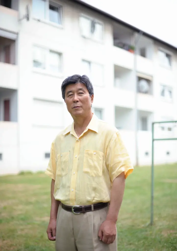 Portrait von Kim CS in einem gelben kurzärmeligen Hemd vor einem hellen Wohngebäude.