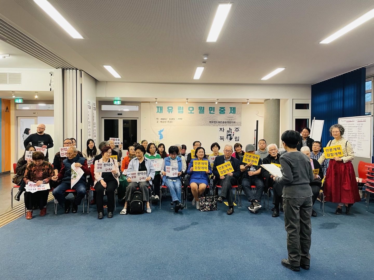 [Ein Foto in der Totale, die eine Anzahl der Teilnehmenden am 45.Mai- Seminar in einem Veranstaltungsraum zeigt. Sie sitzen auf Stühlen und halten kleine Schilder in ihren Händen mit diversen Aufschriften wie z.B: “Medienreform- Reform der Staatsanwaltschaft”, “Festnahme des wegen Hochverrats angeklagten Kriminellen Han Deok-su”]