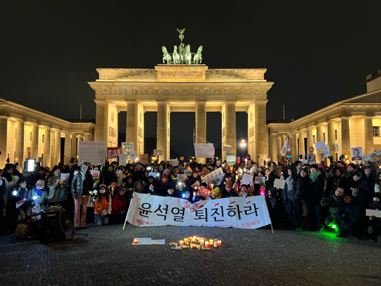 [Das Bild ist ein Gruppenfoto der Demonstrierenden nach der Kundgebung am Abend des 13. Dezember 2024. Die Gruppe steht vor dem beleuchteten Brandenburger Tor. Das große Banner vor der Gruppe sagt “윤석열 즉각 퇴진하라, Yoon Suk-yeol, tritt sofort zurück”]