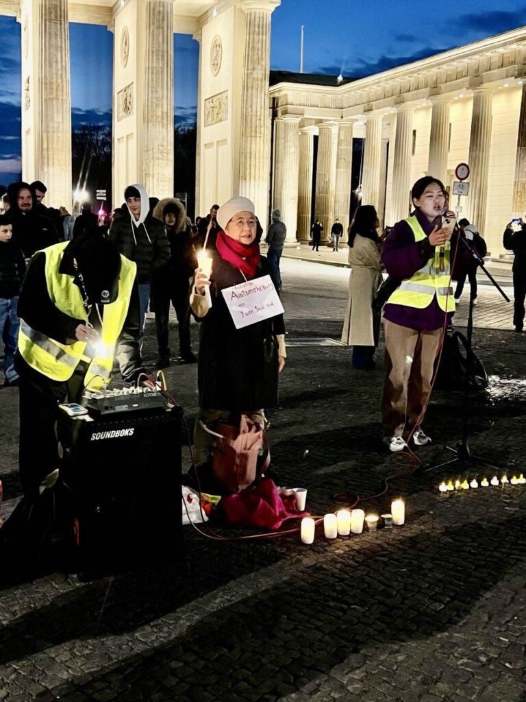 [Das Bild zeigt Seo Eui Ok und Im Dahye bei einer Abendkundgebung. Seo Eui Ok hält eine Kerze und hat ein Poster um den Hals gehängt, das die sofortige Amtsenthebung Yoon Suk-yeols fordert. Dahye steht währenddessen am Mikrophon und moderiert die Kundgebung.]
