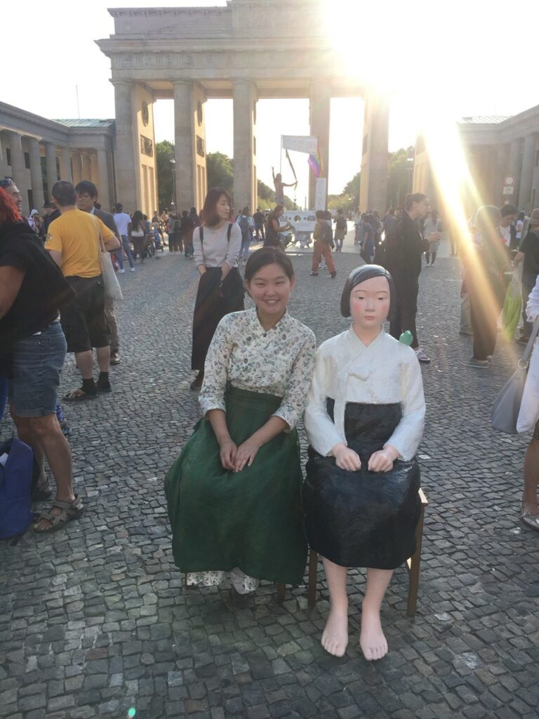 [Das Foto zeigt Dahye mit der transportierbaren Friedensstaute “Yong-i”. Dahye selbst trägt wie die Friedensstaue einen Hanbok mit geblümter Bluse und grünem Rock.]