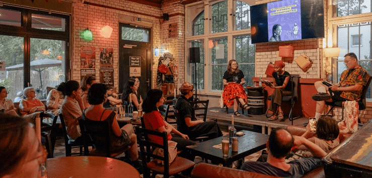 [Jana Schäfer, Fatin Abbas und Timm Young Shik Therre (l. nach r.) sitzen im Café der Kulturfabrik Moabit. Eine Reihe Gäste sitzt vor ihnen.]
