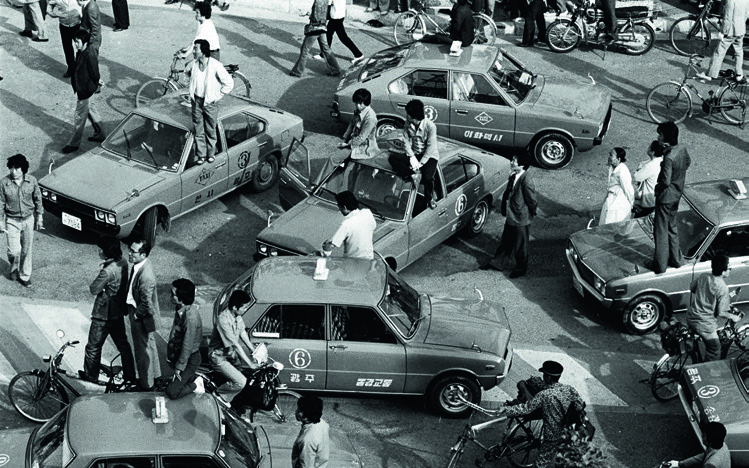 Taxis stehen kreuz und quer auf einer Kreuzung. Junge Männer sitzen auf den Dächern, Bürger*innen stehen auf der Straße und beobachten die Geschehnisse.